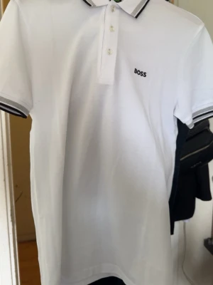 Vit pikétröja från BOSS - Klassisk vit pikétröja från BOSS med svart logga på bröstet. Tröjan har krage och knappar framtill samt svarta detaljer på krage och ärmslut. Perfekt till en stilren och avslappnad look. En jätte ny tröja storlek M!