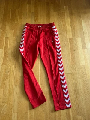 Röda Hummel mjukisbyxor /trackpants - Snygga röda mjukisbyxor från Hummel med vita chevronränder längs sidorna och elastisk midja med dragsko. Byxorna har två fickor och Hummels logga broderad på ena benet. Perfekta för chill eller träning.