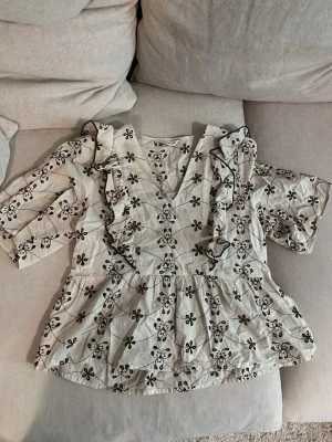 Vit blus med volanger och blommönster - Söt vit blus med brunt blommönster och volangdetaljer längs den V-formade ringningen. Blusen har trekvartsärm och är tillverkad i ett lätt tyg, perfekt för en trendig och avslappnad look. Passar perfekt till jeans eller kjol för en fräsch stil. Köpt på vero moda.