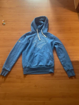 Blå Superdry hoodie - Säljer en blå hoodie från Superdry med stort tryck på bröstet och ficka framtill. Hoodien har vita dragsnören i huvan och ribbade muddar vid ärmar och midja. Perfekt för dig som gillar streetwear och vill ha en skön och snygg tröja.