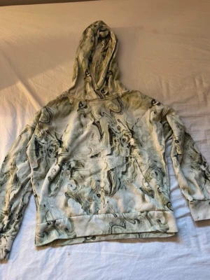 Mönstrad hoodie från Lindex för barn - Säljer en unik hoodie från Lindex med grönvit marmorerad design. Hoodien har stor ficka framtill och en klassisk huva. Tillverkad i mjukt material som är skönt att ha på sig. Perfekt för dig som gillar coola mönster och streetwear vibes.