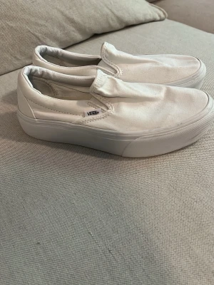 Vita slip-on sneakers från Vans - Klassiska vita slip-on sneakers från Vans med canvasöverdel och tjock vit sula. Minimalistisk design utan snörning och diskret Vans-logga på sidan. Perfekta för en clean och avslappnad stil.