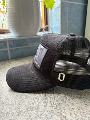 Svart truckerkeps från Onecap - Säljer en svart truckerkeps från Onecap med mesh på sidorna och baksidan för extra ventilation. Framtill finns en stor patch med svartvitt motiv. Kepsen har böjd skärm och justerbar passform. Perfekt för dig som gillar streetwear och unik design.