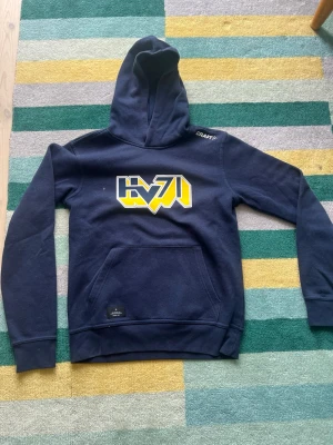 Mörkblå HV71 hoodie från Craft - Snygg mörkblå hoodie från Craft med HV71-logga i gult och vitt på bröstet. Klassisk känguruficka framtill och huva. Perfekt för dig som gillar hockey och vill ha en bekväm och stilren tröja. Tillverkad i mjukt material.