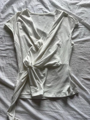 Vit omlottopp från SHEIN - Snygg vit omlottopp från SHEIN med korta ärmar och knyt i sidan. Toppen har v-ringning och är tillverkad i ett mjukt, lätt material som känns skönt mot huden. Perfekt att styla med jeans eller kjol för en fräsch look.