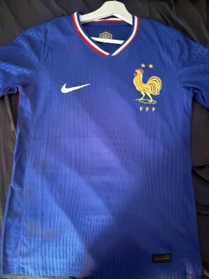 Frankrike fotbollströja Nike blå - Säljer en blå officiell Frankrike fotbollströja från Nike med FFF-logga och tupp på bröstet. Tröjan har vita och röda detaljer i kragen och är tillverkad i ett lätt, ventilerande material. 