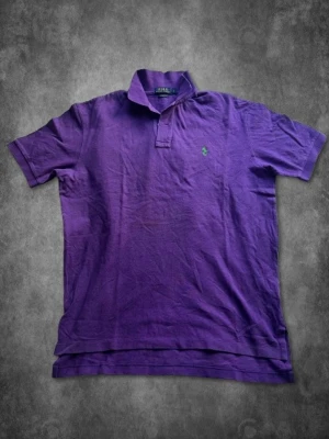Lila pikétröja från Polo Ralph Lauren - Snygg lila pikétröja från Polo Ralph Lauren i klassisk passform. Tröjan har korta ärmar, krage och knappslå framtill samt den ikoniska gröna loggan broderad på bröstet. Perfekt för en avslappnad men stilren look.