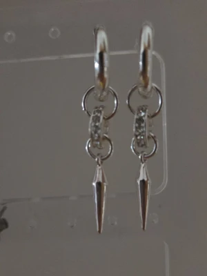 Silvriga örhängen med spetsig detalj - Snygga örhängen i silverfärg med runda ringar och gnistrande stenar på mitten. Hängande spetsig detalj ger en edgy vibe. Perfekta för dig som gillar att sticka ut med accessoarer. Materialet ser ut att vara metall med glittrande stenar.