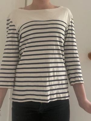 Randig topp med trekvartsärm - Säljer en vit och marinblå randig topp med trekvartsärm och bred halsringning. Tillverkad i mjuk bomull som känns skön mot huden. Perfekt för en avslappnad och stilren look.