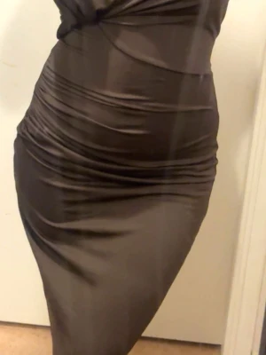 Brun lång klänning  - Elegant mörkbrun åtsittande passform och draperade detaljer längs sidorna. Klänningen har en smickrande siluett och passar perfekt för dig som vill ha en stilren och trendig look. Öppen på ryggen.