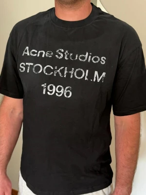 Svart Acne Studios t-shirt med tryck - Cool svart t-shirt från Acne Studios med texten 'Acne Studios STOCKHOLM 1996' i vitt framtill. Klassisk passform med rund hals och korta ärmar. Perfekt för en stilren och trendig look.