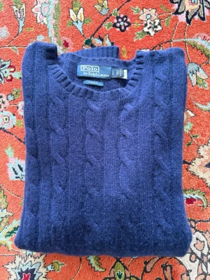 Ralph Lauren kabelstickad cashmeretröja - Säljer en mörkblå kabelstickad tröja från Polo Ralph Lauren i mjuk cashmere. Tröjan har rund halsringning och ribbade muddar. Nypris ligger på 7200kr