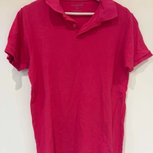 Rosa pikétröja från Primark - En klassisk rosa pikétröja från Primark med korta ärmar och krage. Tröjan har två knappar vid halsen och är tillverkad i mjuk bomull. Perfekt för en avslappnad och stilren look.