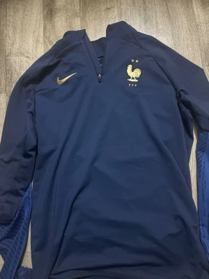 Blå Nike FFF träningströja - Mörkblå träningströja från Nike med halv dragkedja och FFF-logga i guld på bröstet. Tillverkad i Dri-FIT-material som andas, perfekt för fotbollsträning. Guldiga detaljer och diskret mönster på ärmarna ger en sportig look.
