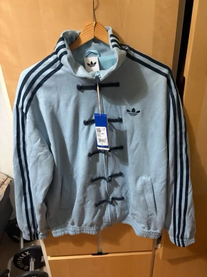 Adidas chinese lunar jacka - Ljusblå fleecejacka från Adidas med svarta snörningsdetaljer framtill och klassiska tre ränder längs ärmarna. Jackan har hög krage, sidofickor och Adidas logga broderad på bröstet. Perfekt för dig som gillar sportig och unik stil.