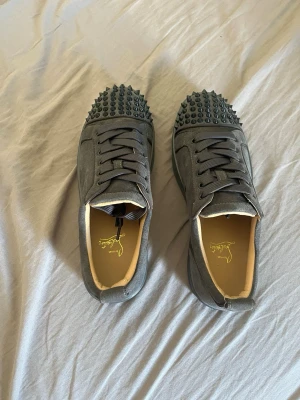 Gråa Louboutin sneakers med nitar - Säljer ett par gråa sneakers från Christian Louboutin i mocka med svarta nitar på tån. Skorna har klassisk låg modell, breda grå skosnören och svart sula. Insidan är ljusbeige och märket syns på innersulan. Perfekta för dig som vill sticka ut med en edgy stil.