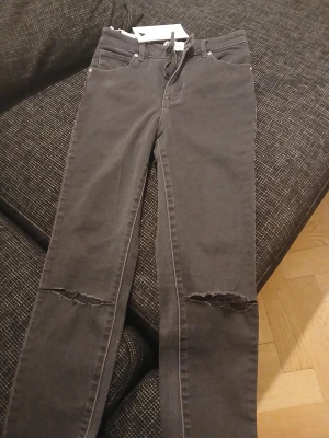 Svarta ripped jeans från Gina Tricot - Snygga svarta jeans från Gina Tricot med raka ben och slitningar över knäna. Klassisk femficksmodell med bälteshällor och knappgylf. Jeansen har en cool streetstil och är tillverkade i mjukt denimtyg.