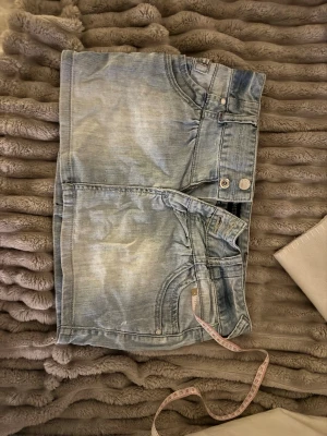 Ljusblå jeanskjol med fickor - Säljer en ljusblå jeanskjol i kort modell med klassiska fem fickor, bälteshällor och knappgylf. Kjolen har slitna detaljer och en avslappnad look, perfekt för dig som gillar denim. Passar till många olika stilar!