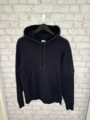 C.P. Company Hoodie - CP Company hoodie i mörkblå färg. Riktigt clean och stilren modell med klassiska lens-detaljen på armen. Väldigt fint skick och skön passform.