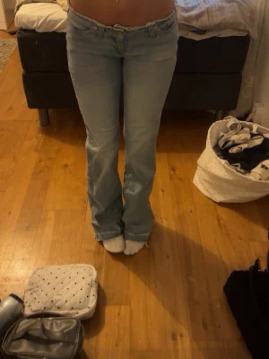 Ljusblå bootcut jeans - Säljer ett par ljusblå bootcut jeans med låg midja och klassisk femficksdesign. Jeansen har en snygg utsvängd passform nertill och är perfekta för en avslappnad och trendig look. Materialet är denim i bomull med lite stretch för extra komfort. Jeansen har ett litet hål där framme men går att sy