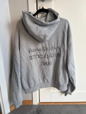 Grå hoodie från Acne Studios - Snygg grå hoodie från Acne Studios med tryck på ryggen där det står 'Acne Studios STOCKHOLM 1996'. Hoodien har huva, långa ärmar och ribbade muddar. Perfekt för en avslappnad och trendig look.