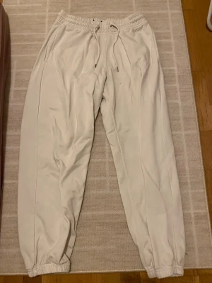 Beiga mjukisbyxor med dragsko - Säljer ett par beiga mjukisbyxor med resår i midjan och dragsko. Byxorna har fickor på sidorna och resår vid bensluten för en skön och avslappnad look. Perfekta för chill dagar eller när du vill vara bekväm.