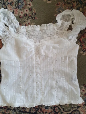 Vit spetsblus med knappar - Söt vit blus med broderade detaljer och knappar framtill. Blusen har rynkad smock baktill, volangkant och romantiska puffade axelband med spets. Materialet är lätt och luftigt, perfekt för sommarens outfits.