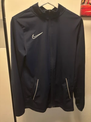 Nike mörkblå tränings tröja med dragkedja - Mörkblå tränings tröja från Nike med dragkedja, hög krage och två fickor med vita detaljer. Jackan har lång ärm och är tillverkad i ett lätt, syntetiskt material som passar perfekt till sport och träning. Funkar även utmärkt att matcha med jeans eller shorts. Klassisk Nike-logga på bröstet.