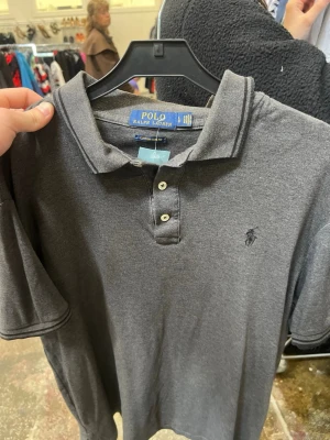 Grå pikétröja från Polo Ralph Lauren - Klassisk grå pikétröja från Polo Ralph Lauren med krage, knappar framtill och broderad logga på bröstet. Tröjan har korta ärmar och är tillverkad i mjuk bomull för en bekväm känsla.