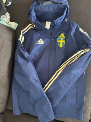 Blå Adidas Sverige vindjacka (passar xs/s) - Snygg blå vindjacka från Adidas med Sveriges landslagsemblem på bröstet och vita ränder längs ärmarna. Jackan har huva, dragkedja framtill och är tillverkad i lätt, vindtätt material. Perfekt för dig som vill visa ditt stöd för Sverige.