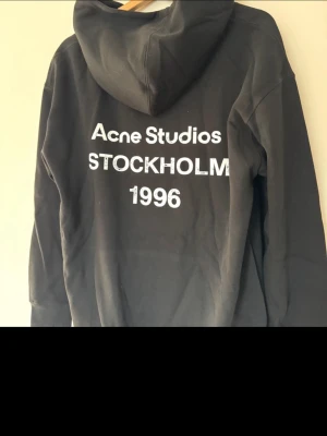 Svart hoodie Acne Studios Stockholm - Svart hoodie från Acne Studios med vit text på ryggen: 'Acne Studios STOCKHOLM 1996'. Klassisk design med huva och långa ärmar. Perfekt för dig som gillar stilrena och trendiga plagg. Materialet ser ut att vara mjukt och bekvämt, troligen bomull. Storleken är Xl men passar bättre på L/M