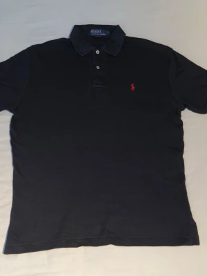 Svart pikétröja från Ralph Lauren - Klassisk svart pikétröja från Ralph Lauren med röd broderad logga på bröstet. Tröjan har krage, två knappar framtill och är tillverkad i mjuk bomull. Perfekt för en stilren och avslappnad look.