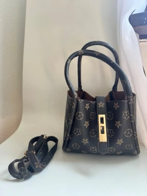 Brun handväska med monogram och axelrem NY - Snygg brun handväska  med klassiskt monogrammönster och guldfärgat spänne. Väskan har två handtag och avtagbar axelrem för flexibel användning. Tillverkad i tåligt lädermaterial och har en kompakt, boxig form.