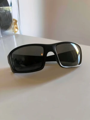 Svarta sportiga solglasögon  - Snygga svarta solglasögon med bred båge och rektangulära, mörka linser. Glasögonen har en sportig design och passar perfekt för dig som gillar en cool och modern look. Tillverkade i syntetmaterial för en lätt och bekväm känsla.