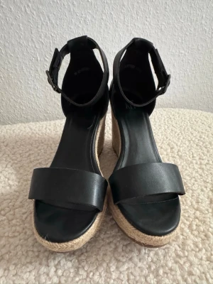Svarta kilklackade espadrillos med rem som nya. - Snygga svarta sandaler med bred rem över tårna och justerbart spänne vid ankeln. Skorna har en hög kilklack i flätad jute som ger en somrig känsla. Perfekta till jeans eller klänning för en trendig look. Använd 2 gånger.