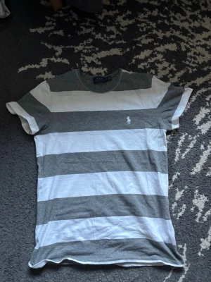 Randig t-shirt från Polo Ralph Lauren - Snygg t-shirt från Polo Ralph Lauren med breda horisontella ränder i grått och vitt. Liten i storlek, passar XS, med Ikoniska broderade loggan på bröstet. Perfekt till sommaren