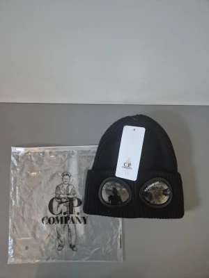 C.P. Company mössa - Svart ribbstickad mössa från C.P. Company med ikoniska goggles framtill. Tillverkad i mjukt material som håller värmen och ger en cool streetwear-look. Perfekt accessoar för dig som vill sticka ut och addera en unik touch till din outfit.