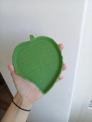 Grön bricka formad som ett löv - Säljer en liten grön bricka i plast formad som ett löv. Perfekt för småsaker, smycken eller dekoration. Brickan har tydliga ådror på ovansidan och en liten stjälkdetalj. Enkel och stilren design som sticker ut.