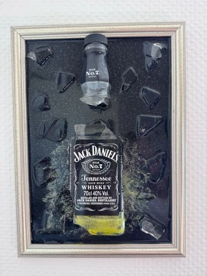 Jack Daniel's tavla  - Unik tavla med en Jack Daniel's-flaska monterade på svart bakgrund, omgivna av krossade glasbitar. Silverfärgad ram och tydlig etikett på flaskan. Perfekt som dekoration för hemmabaren eller som present till whiskeyälskare. Tillverkad av mig själv 