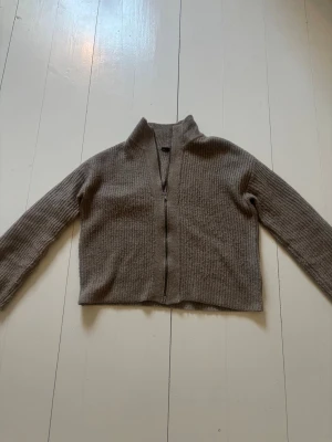 Beige ribbstickad ziptröja 157 - Beige ribbstickad tröja från 157 med dragkedja framtill och hög krage. Tröjan har långa ärmar och en avslappnad passform, perfekt för lager-på-lager. Materialet är mjukt och ger en skön känsla mot huden.
