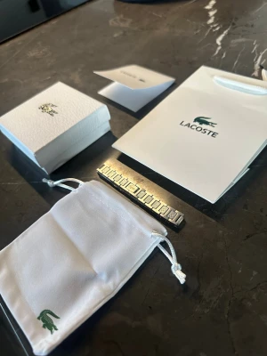 Silverfärgat armband från Lacoste - Snyggt silverfärgat armband från Lacoste med rektangulära länkar och diskreta detaljer. Armbandet kommer med originalförpackning, tygpåse och presentpåse, allt med Lacoste-loggan. Perfekt accessoar för dig som gillar stilrena smycken.