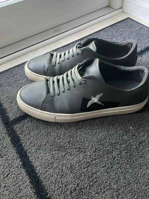 Svarta Arigato sneakers i läder - Snygga grå/svarta sneakers från Axel Arigato i slätt läder med vit sula och ikonisk fågelbrodyr på sidan. Klassisk låg modell med matchande svarta skosnören och diskret logga på hälen. Perfekta för en clean och stilren look dem sitter även som 45 för dem som har den storleken.