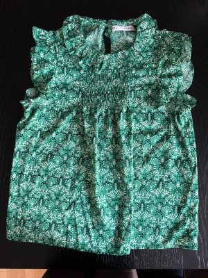Gullig grön blommig blus med volangärmar - Snygg grön blus med vitt och svart blommönster och volangdetaljer vid axlar och hals. Smockad upptill och rundad krage med liten knapp i nacken. Ärmlös modell i lätt och luftig kvalitet, perfekt för sommardagar. Varan är i mycket bra skick!💚