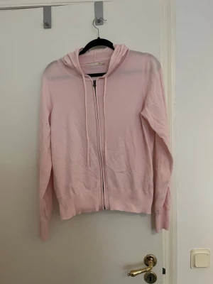 Ljusrosa hoodie med dragkedja - Ljusrosa hoodie med dragkedja framtill och huva med dragsko. Tillverkad i ett mjukt och tunt trikåmaterial. Långa ärmar och ribbade muddar vid ärmslut och nederkant.