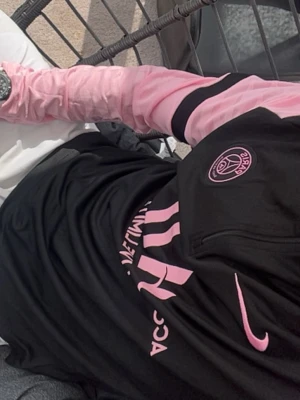 PSG träningströja svart/rosa Nike - Snygg långärmad träningströja från Paris Saint-Germain i svart med rosa detaljer och logga. Tröjan har halv dragkedja framtill, rosa ärmar med svarta ränder och PSG-emblem på bröstet. Perfekt för fotbollsträning eller sportiga outfits.