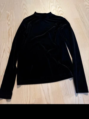 Svart polotopp i sammet - Snygg svart polotopp i mjuk sammet med lång ärm. Perfekt för dig som gillar en stilren och elegant look. Polokrage och figurnära passform ger en trendig vibe.