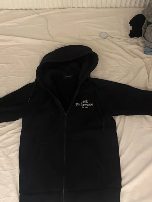 Svart Peak Performance hoodie med dragkedja - Svart hoodie från Peak Performance med vit broderad logga på bröstet. Modellen har dragkedja framtill, huva med dragsko och mjukt fleecefoder. Perfekt för en chill och sportig stil.