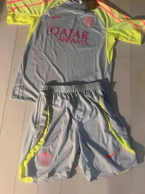 PSG träningsset grå/gul Nike - Paris Saint-Germain träningsset med t-shirt och shorts i ljusgrått med neon-gula och rosa detaljer. PSG-logga och Qatar Airways-tryck på tröjan, Nike-logga på både tröja och shorts. Tillverkat i lätt och ventilerande funktionsmaterial, perfekt för fotbollsträning.