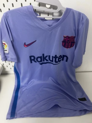 Lila FC Barcelona t-shirt Nike - Lila FC Barcelona t-shirt från Nike med klubbmärke och Rakuten-tryck på bröstet. Kortärmad modell i lätt polyester, v-ringad hals och La Liga-märke på ärmen. Snygga blå detaljer längs sidan och UNICEF-tryck på ryggen. Perfekt för fotbollsfans som vill sticka ut.