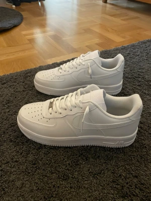 Nike Air Force 1 vita sneakers - Klassiska helvita Nike Air Force 1 sneakers i lågt utförande. Skorna har ovandel i slätt läder, perforeringar på tån för ventilation och den ikoniska swoosh-loggan på sidan. Tjock sula med AIR-dämpning och vita skosnören. Perfekta för en clean och stilren look.